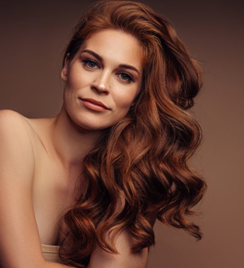 A confident woman with long wavy hair and a casual pose, captured in a studio setting against a warm-toned background. Her subtle expression and relaxed posture make for a captivating portrait. This image conveys elegance, sophistication, and natural beauty, ideal for themes like fashion, beauty, or personal care advertising.